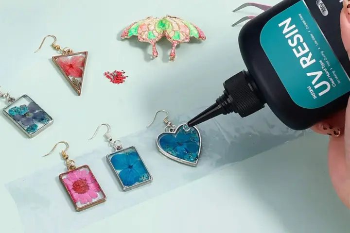 A hand applies UV resin to a heart-shaped earring with blue flowers, demonstrating how UV resin works to create beautiful jewelry.