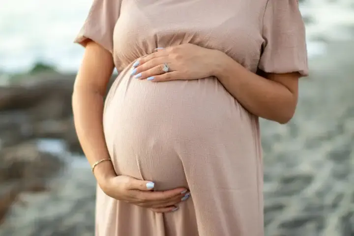 Brzuszek ciężarnej kobiety w beżowej sukience, dłonie delikatnie go obejmują. Warto pamiętać o bezpieczeństwie i unikać stosowania **retinolu w ciąży**.