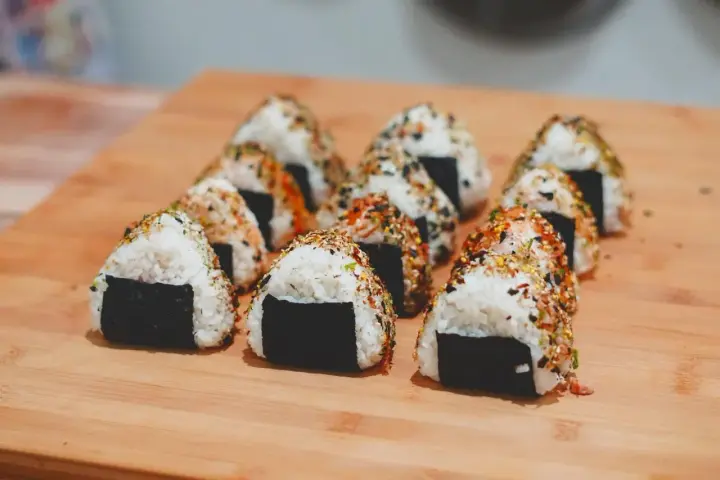 Rząd trójkątnych onigiri z kurczakiem, posypanych sezamem i przyprawami, ułożonych na drewnianej desce.