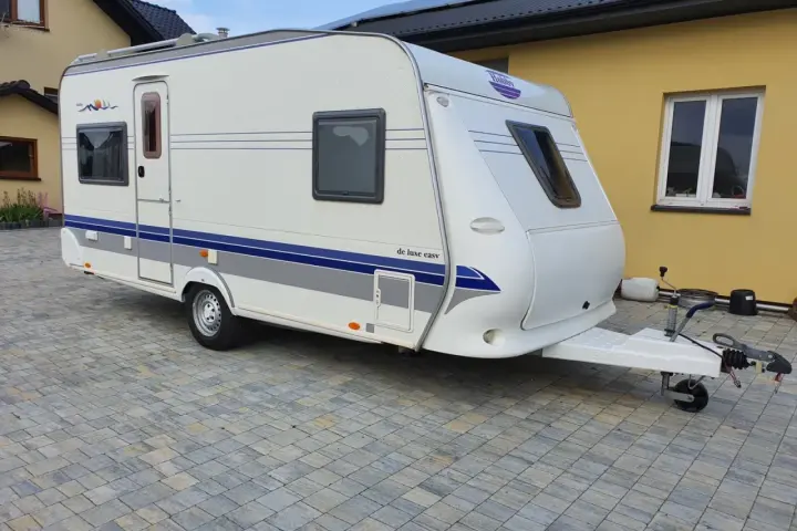 Biała przyczepa kempingowa Hobby 460 UFE "de luxe easy" stoi na kostce brukowej przed żółtym budynkiem.