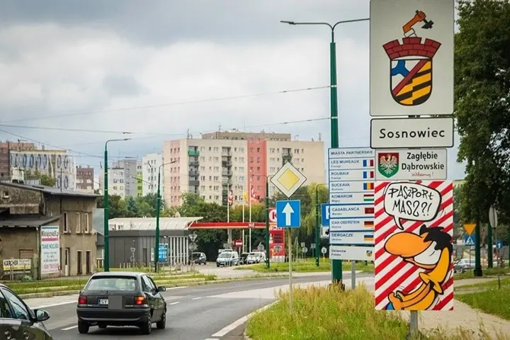 Znak drogowy "Sosnowiec" i humorystyczny napis "Paszport masz?!" przy wjeździe do miasta. W tle bloki mieszkalne.