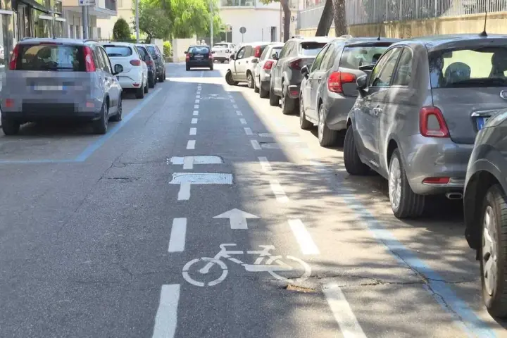 Strada a senso unico con corsia ciclabile delimitata da strisce bianche e blu. Auto parcheggiate lungo il lato destro.