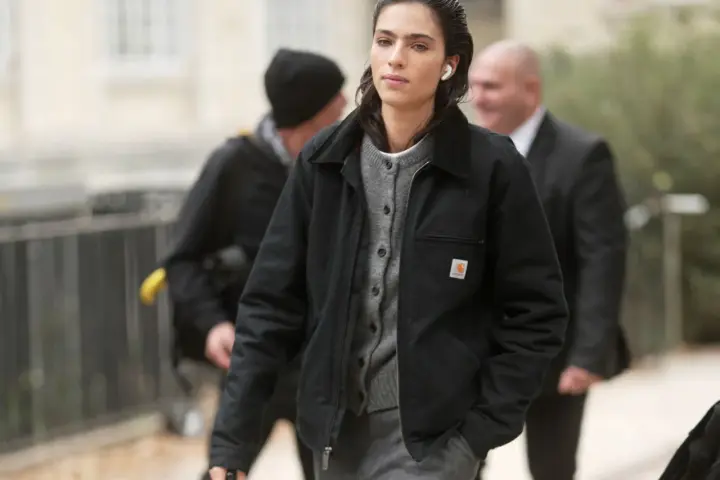 Donna con giacca Carhartt nera e cardigan grigio, cuffiette bianche, cammina con sicurezza.