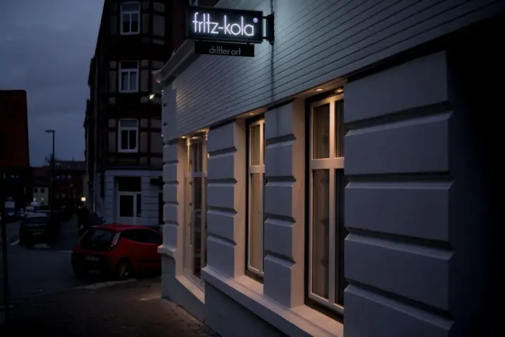 Neon-Schild "fritz-kola" über dem Eingang "dritter ort kiel". Ein rotes Auto parkt auf der Straße.