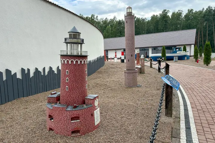 Miniatury latarni morskich na żwirowym placu, z budynkiem i łodzią w tle.