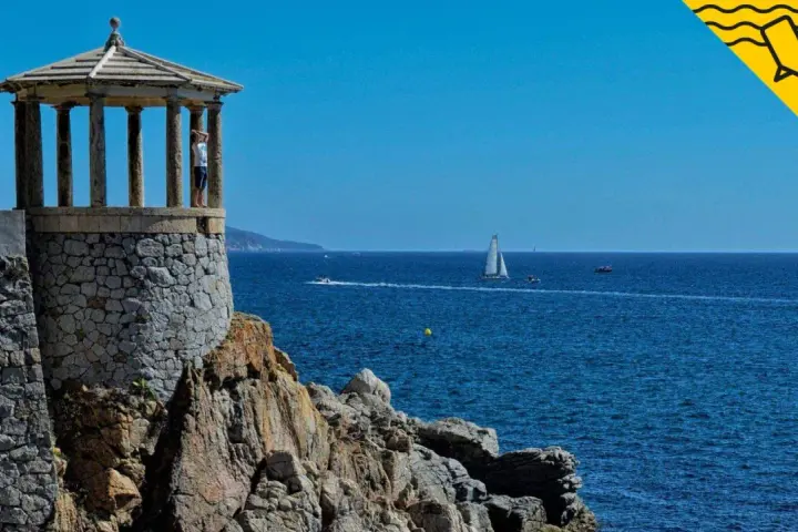 Un mirador de piedra con columnas se alza sobre rocas, con el mar azul y veleros al fondo. Un lugar perfecto para pasear por los **caminos de ronda**.