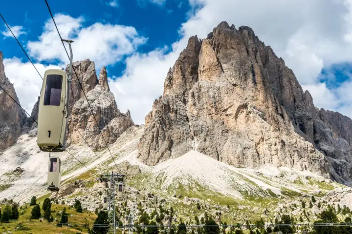 Cabine della funivia salgono verso le imponenti vette del Sassolungo, un anello di roccia maestoso sotto un cielo azzurro con nuvole bianche.