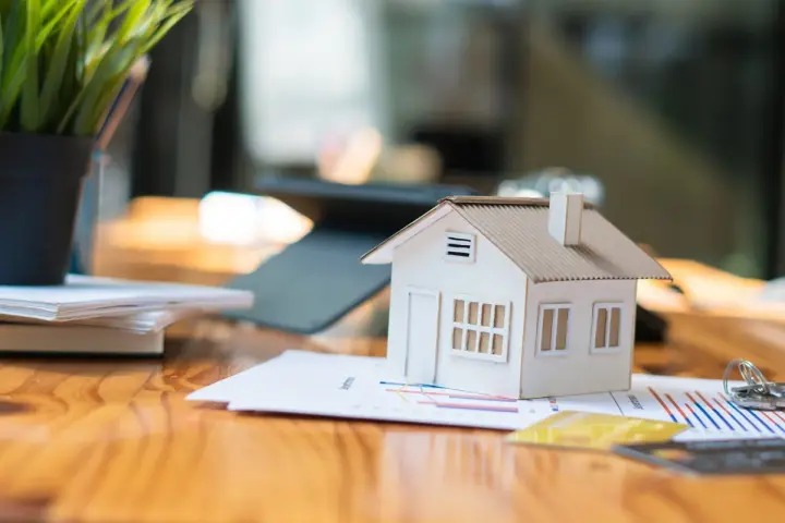 Maqueta de casa sobre gráficos, sugiriendo cómo saber si una casa tiene cargas. Llaves y tarjetas de crédito cerca.