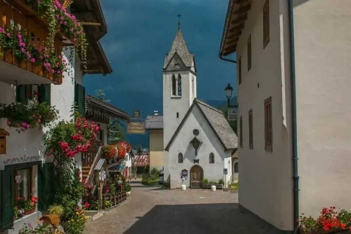 Un pittoresco vicolo a Rocca Pietore, con case fiorite e una chiesa dal cielo tempestoso.