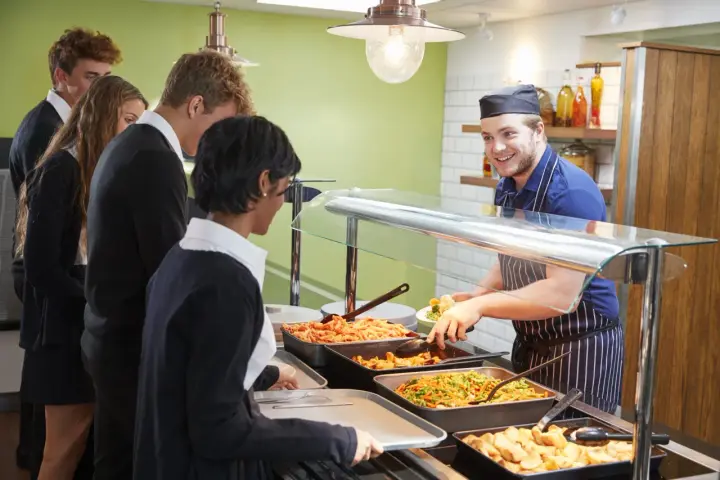 Des élèves font la queue pour le déjeuner à la cantine. Un employé souriant sert des légumes. Un contrat extra restauration est en cours de discussion.