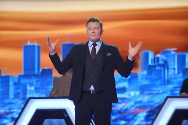 Rafał Brzozowski w garniturze, z rękami uniesionymi na boki, prowadzi program "Jaka to melodia?".