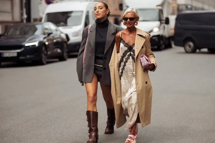 Dos mujeres pasean por la calle. Una lleva un vestido lencero de invierno con un abrigo y la otra un traje gris.