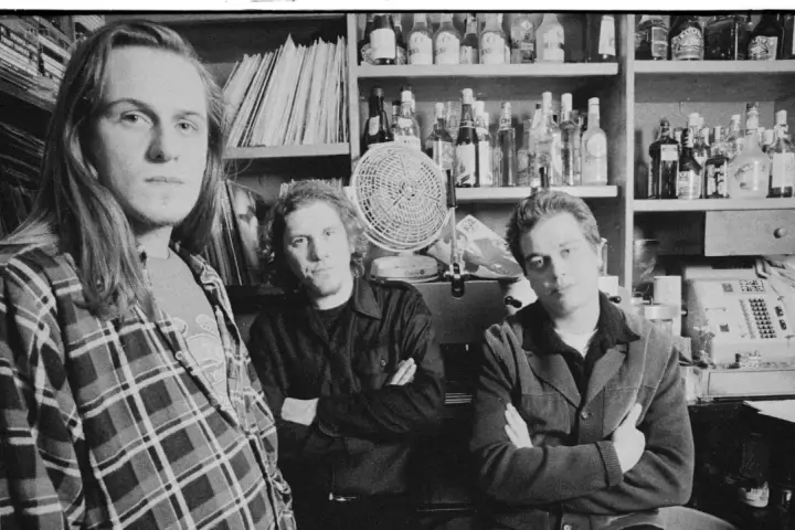 Tres hombres posan en blanco y negro, con estanterías de discos y botellas detrás.