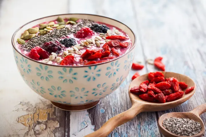 Miseczka smoothie z owocami, nasionami chia, pestkami dyni i słonecznika. Obok łyżki z jagodami goji i nasionami chia. Pyszny sposób, jak jeść nasiona chia.