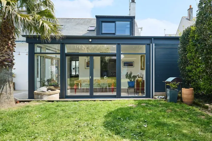 Une extension de maison moderne avec une immense baie vitrée s'ouvrant sur un jardin verdoyant. Une palme se dresse à gauche.