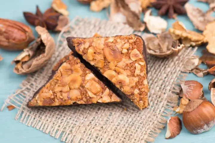 Zwei vegane Nussecken mit Schokoladenrand, bedeckt mit gehackten Nüssen, liegen auf einem Jutesack, umgeben von verschiedenen Nüssen und Sternanis.