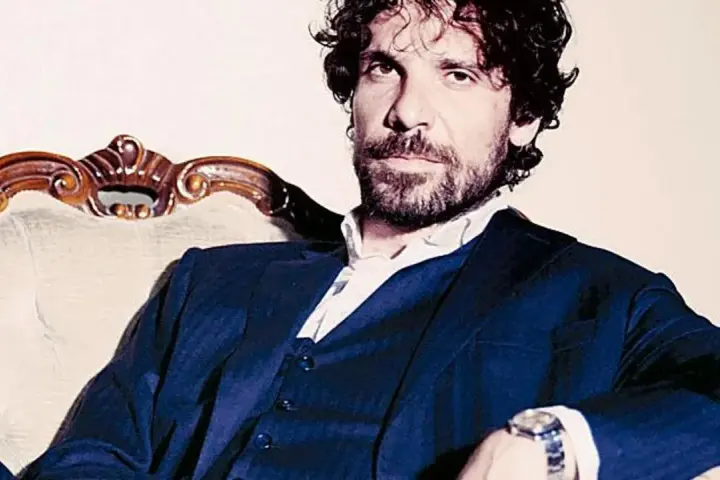 L'attore Montanari, con barba e capelli ricci, indossa un completo blu e una camicia bianca, seduto su una poltrona d'epoca.