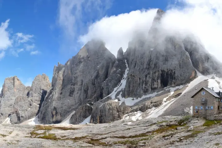 Rifugio in pietra ai piedi di imponenti vette rocciose, con neve residua e cielo azzurro. Un luogo incantevole al passo Valles.