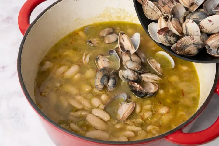 Alubias cremosas con almejas en una olla roja, añadiendo más almejas de una sartén.