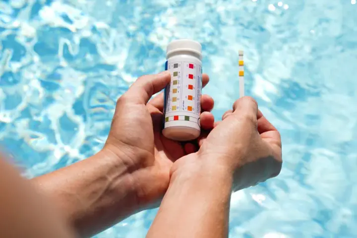 Hands hold a pool test strip and bottle, showing how to make pool water clear by checking chemical levels.