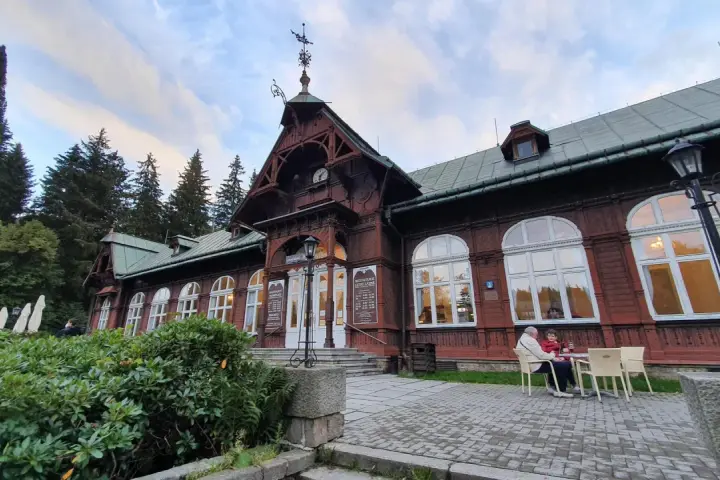Drewniany pawilon z zegarem i karłowata studzienka przed wejściem. Na zewnątrz siedzą ludzie przy stoliku.