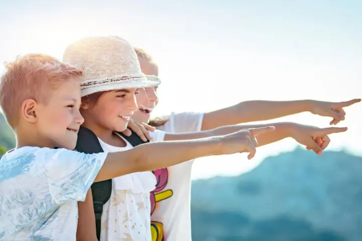 Trois enfants souriants, portant des sacs à dos, pointent du doigt vers la droite, sous un ciel ensoleillé.
