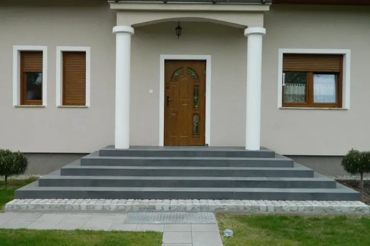 Eleganckie, szare schody zewnętrzne prowadzące do drewnianych drzwi. Świetny pomysł na schody zewnętrzne, podkreślający nowoczesny styl domu.