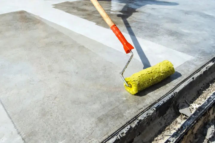 Wałek z żółtym włosiem rozprowadza środek do impregnacji betonu na szarej powierzchni.