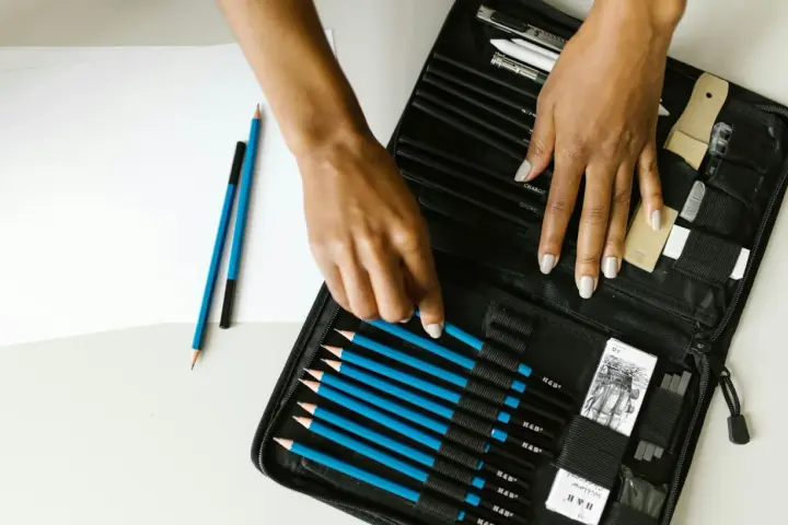Un étui ouvert révèle une sélection de crayons de différents types pour dessiner, avec des mains choisissant un crayon bleu.