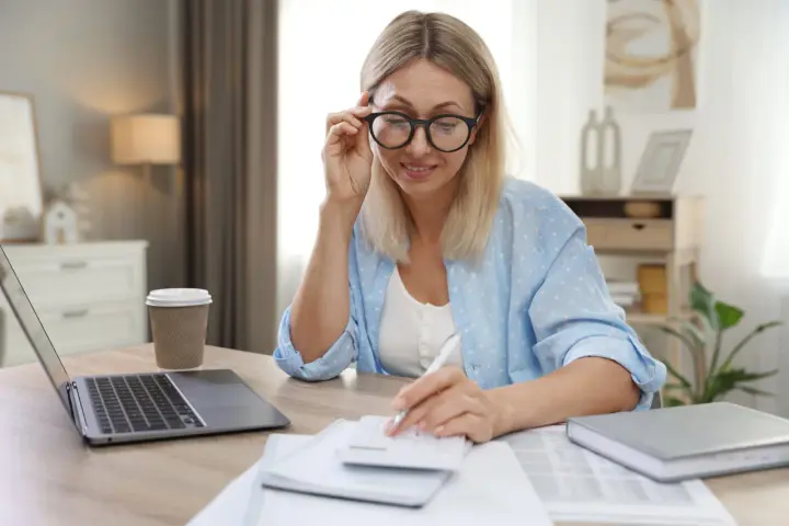 Femme aux lunettes calculant, laptop et café à proximité. Elle semble gérer des finances, peut-être une réduction de capital non motivée par des pertes.