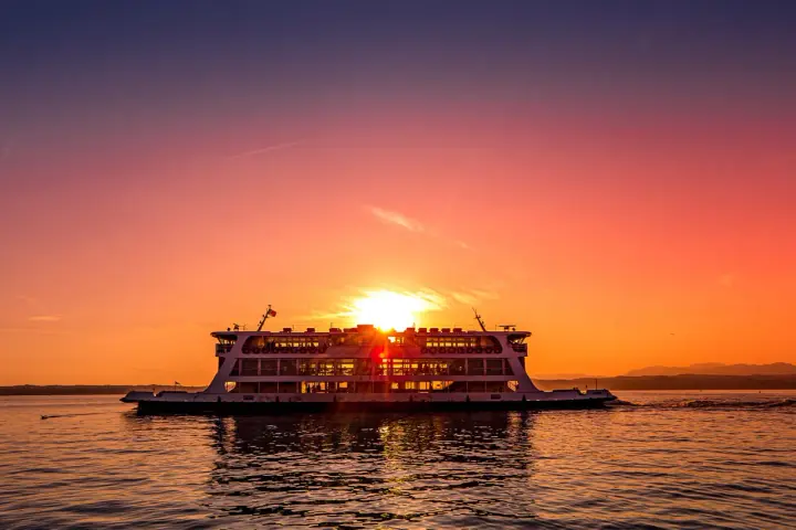 Traghetto in navigazione sul Lago di Garda al tramonto, con il sole che illumina il cielo e il riflesso sull'acqua.