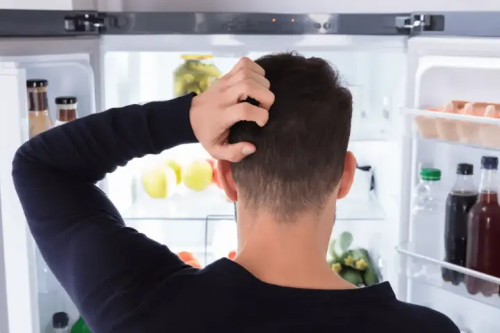 Un homme se gratte la tête devant un réfrigérateur ouvert, plein de nourriture. Il semble réfléchir à un réglage frigo.