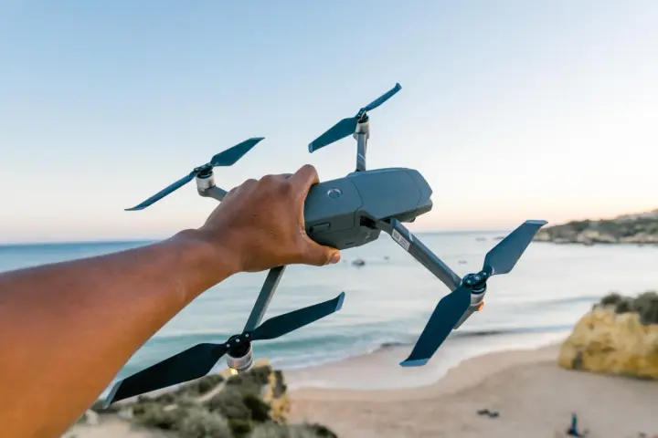 A hand holds a drone over a European beach, ready for flight.