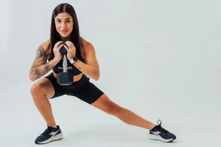 Donna con tatuaggi esegue affondo laterale con manubrio, dimostrando cos'è la palestra funzionale.