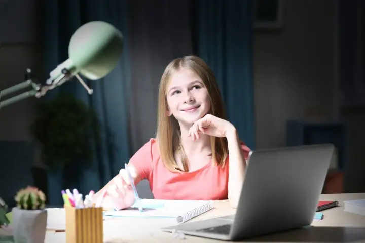 Uśmiechnięta dziewczynka z długopisem w dłoni, zastanawia się nad zadaniem. Obok laptop, dlatego jaka to część mowy?