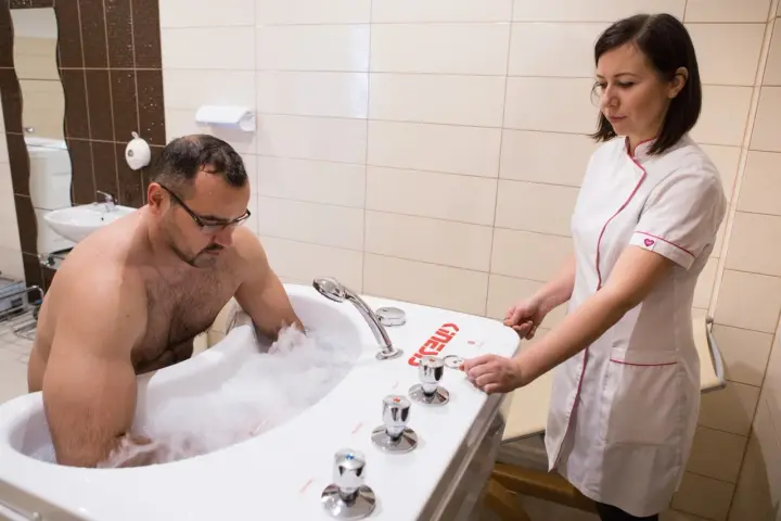 Mężczyzna w wannie z pianą, terapeuta przygotowuje masaż wirowy kończyn dolnych.