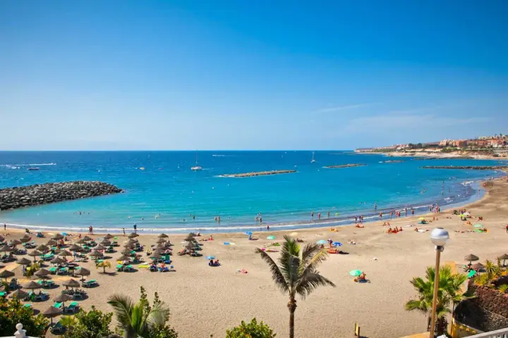 Spiaggia affollata con ombrelloni e lettini, mare azzurro e città costiera sullo sfondo. Sembra l'isola più bella delle Canarie.