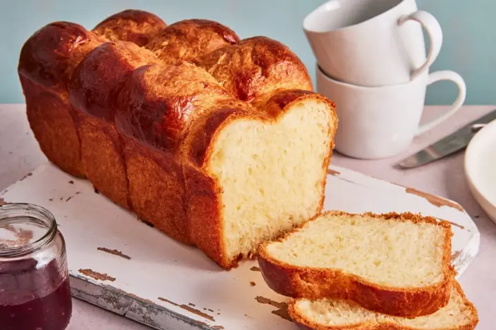 Ein goldbraunes Brioche Brot Rezept, aufgeschnitten auf einem Holzbrett mit Marmelade und Tassen im Hintergrund.