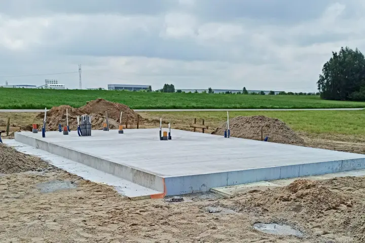 Gotowa płyta fundamentowa na budowie, otoczona piaskiem i zielenią. W tle widać pola i budynki przemysłowe.