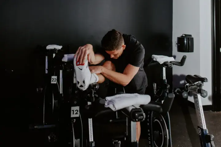 Man in black shirt stretches his outer hamstring by holding his foot, surrounded by stationary bikes in a gym.