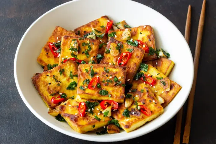 Chrupiące kawałki tofu satay, posypane chili, sezamem i ziołami, podane w białej misce z pałeczkami.