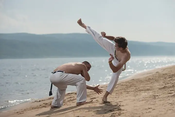 Dwóch mężczyzn ćwiczy capoeirę na plaży. Jeden wykonuje wysokie kopnięcie, drugi jest w pozycji obronnej. Widać ich płynny ginga styl gry.