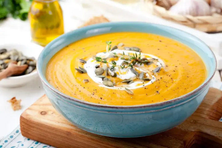 Vellutata di zucca cremosa, guarnita con panna, semi di zucca e timo, servita in una ciotola azzurra su un tagliere di legno.