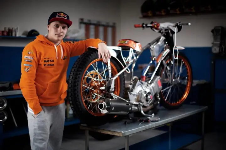 Młody żużlowiec w pomarańczowej bluzie opiera się o swój motocykl żużlowy, gotowy do kolejnego wyścigu.
