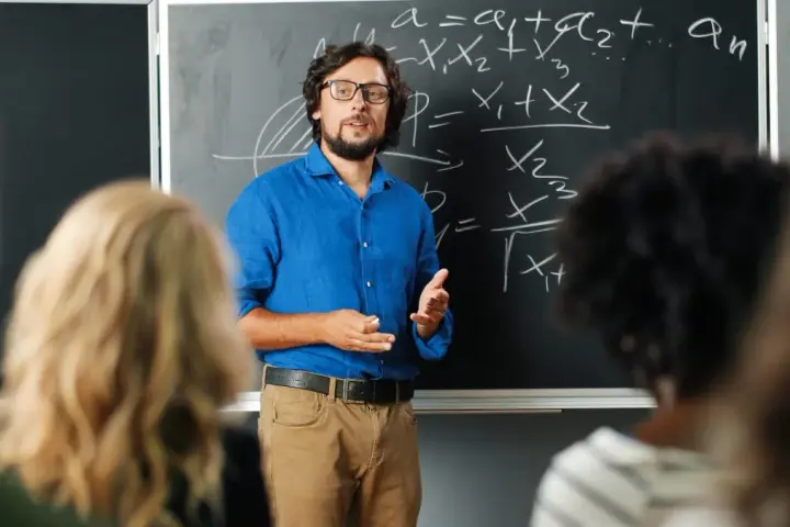 Nauczyciel w niebieskiej koszuli tłumaczy matematykę przy tablicy. Czy nauczyciel jest funkcjonariuszem publicznym? To pytanie nurtuje wielu.