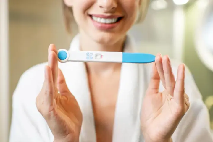 Uśmiechnięta kobieta w szlafroku trzyma test ciążowy z dwoma kreskami. To moment, kiedy warto zastanowić się, kiedy zrobić test ciążowy.