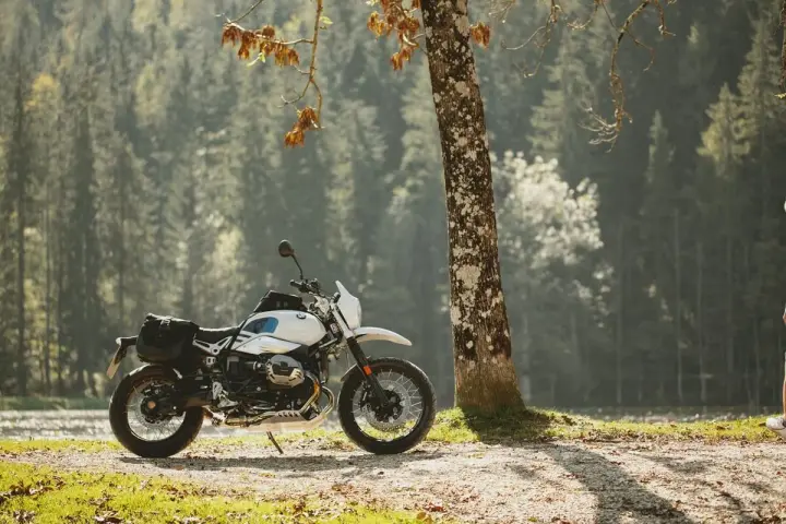 Biały motocykl z sakwami, zaparkowany przy drzewie w lesie. Idealny do testów w rankingu opon motocyklowych.