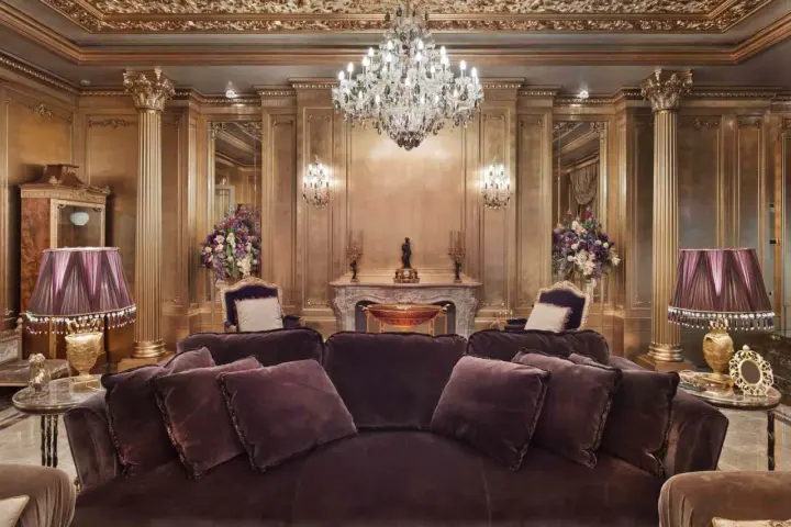 Salón lujoso con sofás de terciopelo morado, lámparas doradas y un candelabro de cristal. El techo y las paredes doradas evocan el estilo barroco español.