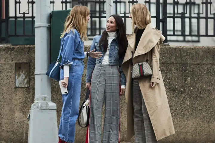 Tres mujeres conversan, una con un traje completo de **denim**, otra con chaqueta **denim** y pantalones a cuadros, y la tercera con un abrigo beige y pantalones a cuadros.