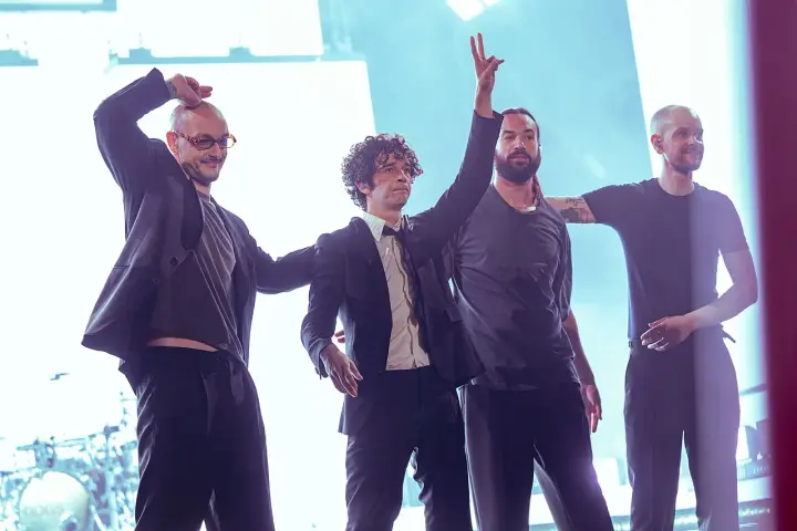 Matty Healy i zespół The 1975 na scenie. Muzycy pozdrawiają publiczność, jeden z nich unosi rękę w geście pokoju.
